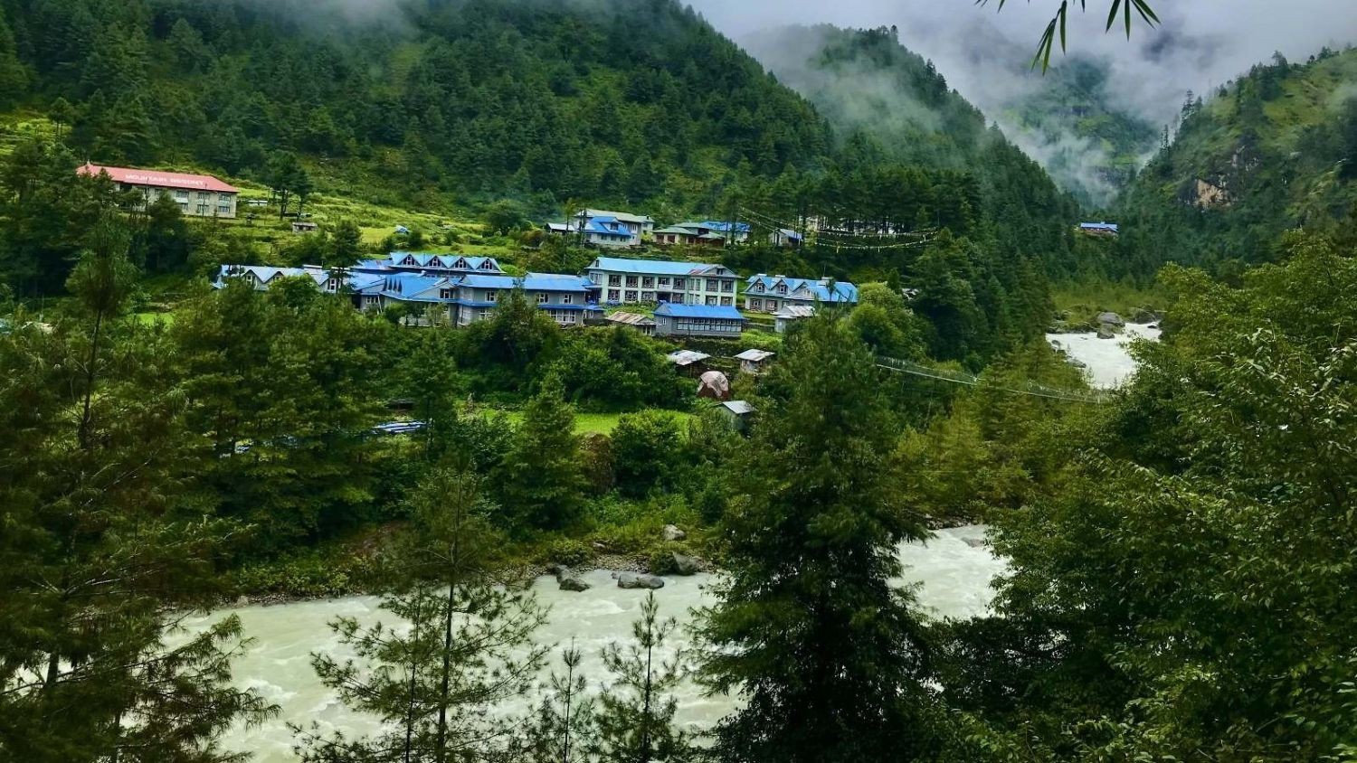 Phakding village in the Everest region with riverside lodges, lush green forests, misty Himalayan hills, and the Dudh Koshi River flowing through the valley