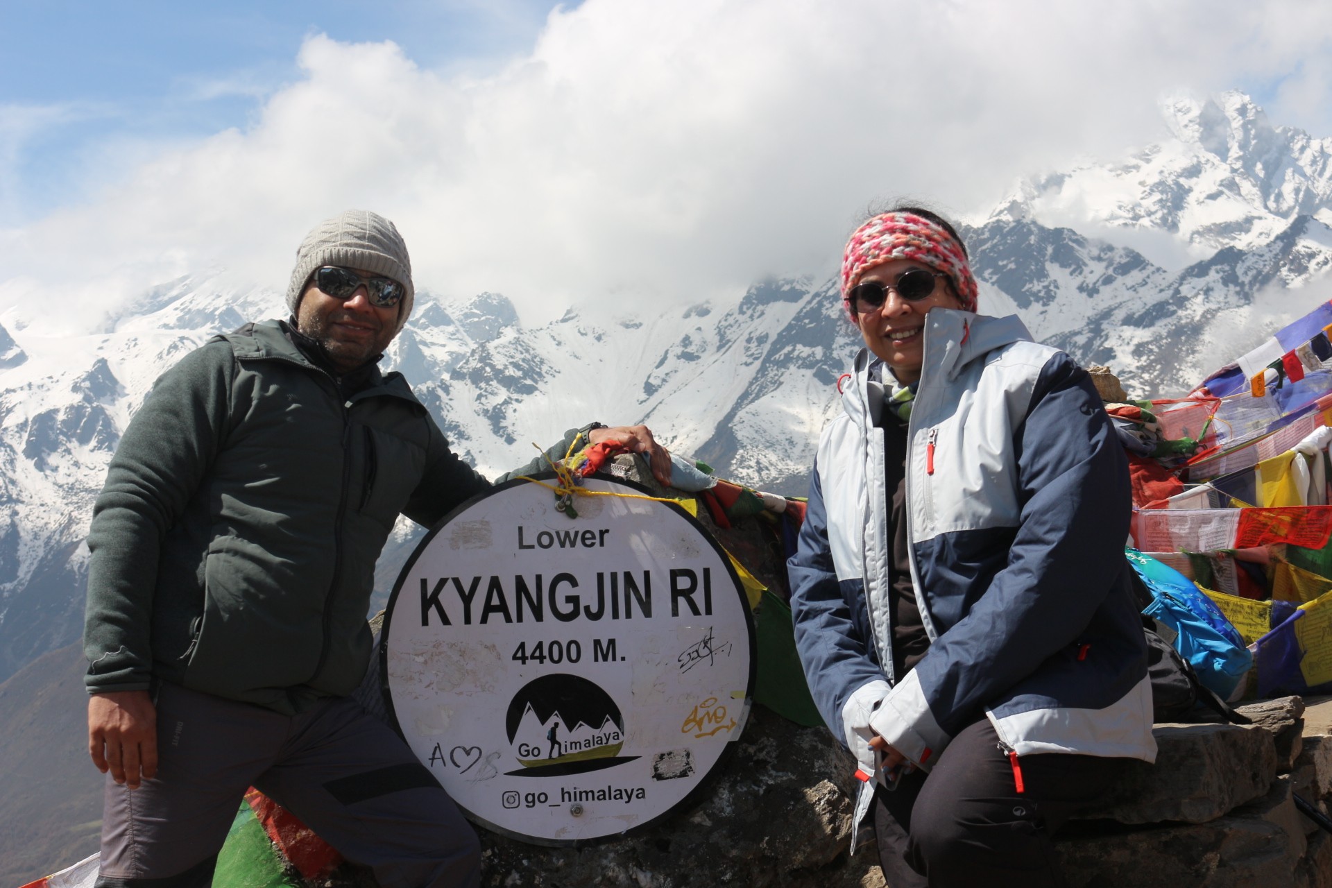 Trekkers reaching the summit of Kyanjin Ri, standing on a rocky viewpoint overlooking the Langtang Himalayan range, glaciers, and vast alpine valleys under a clear blue sky.