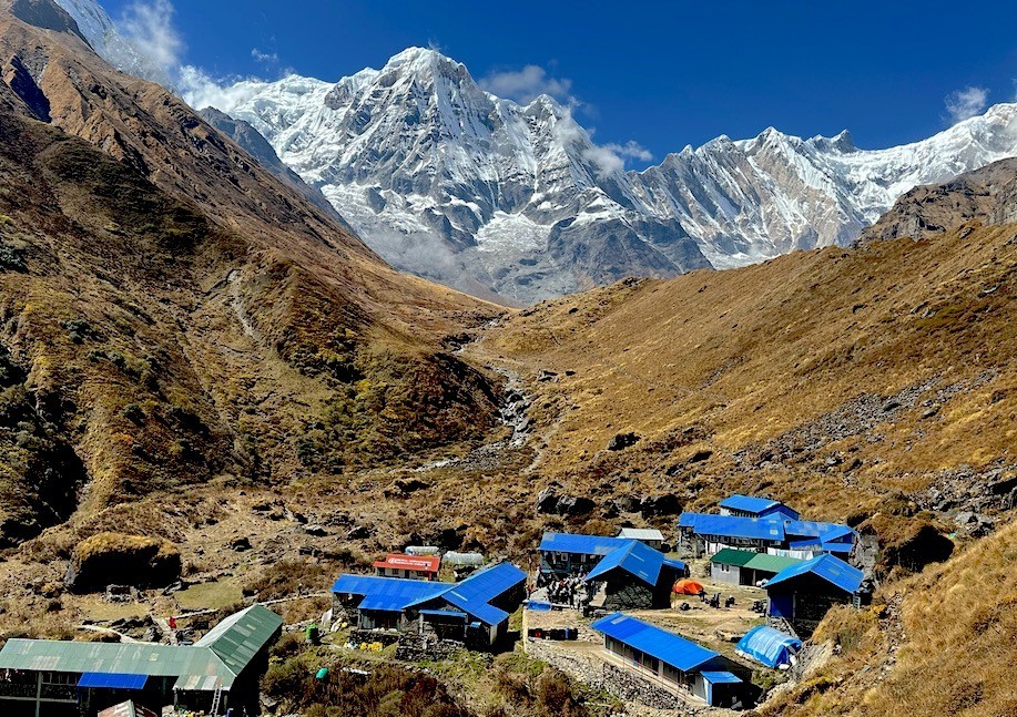Machhapuchhre Base Camp (MBC) with blue-roofed teahouses surrounded by towering snow-covered peaks, including Machhapuchhre (Fishtail) and Annapurna South, on the Annapurna Base Camp Trek in Nepal.