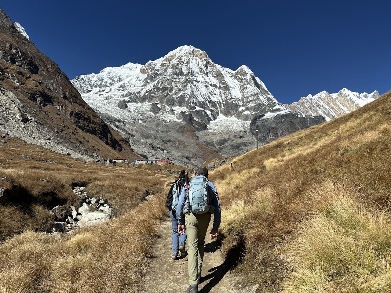 Trekker hiking toward Annapurna Base Camp under clear blue skies with dry trails, sunny weather, and spectacular Himalayan mountain views in November.