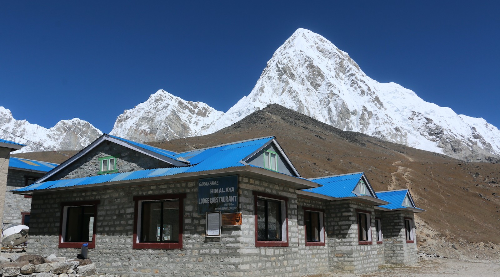 Mountain lodge at Gorak Shep with panoramic view of Pumori and surrounding Himalayan peaks under clear high-altitude sky.