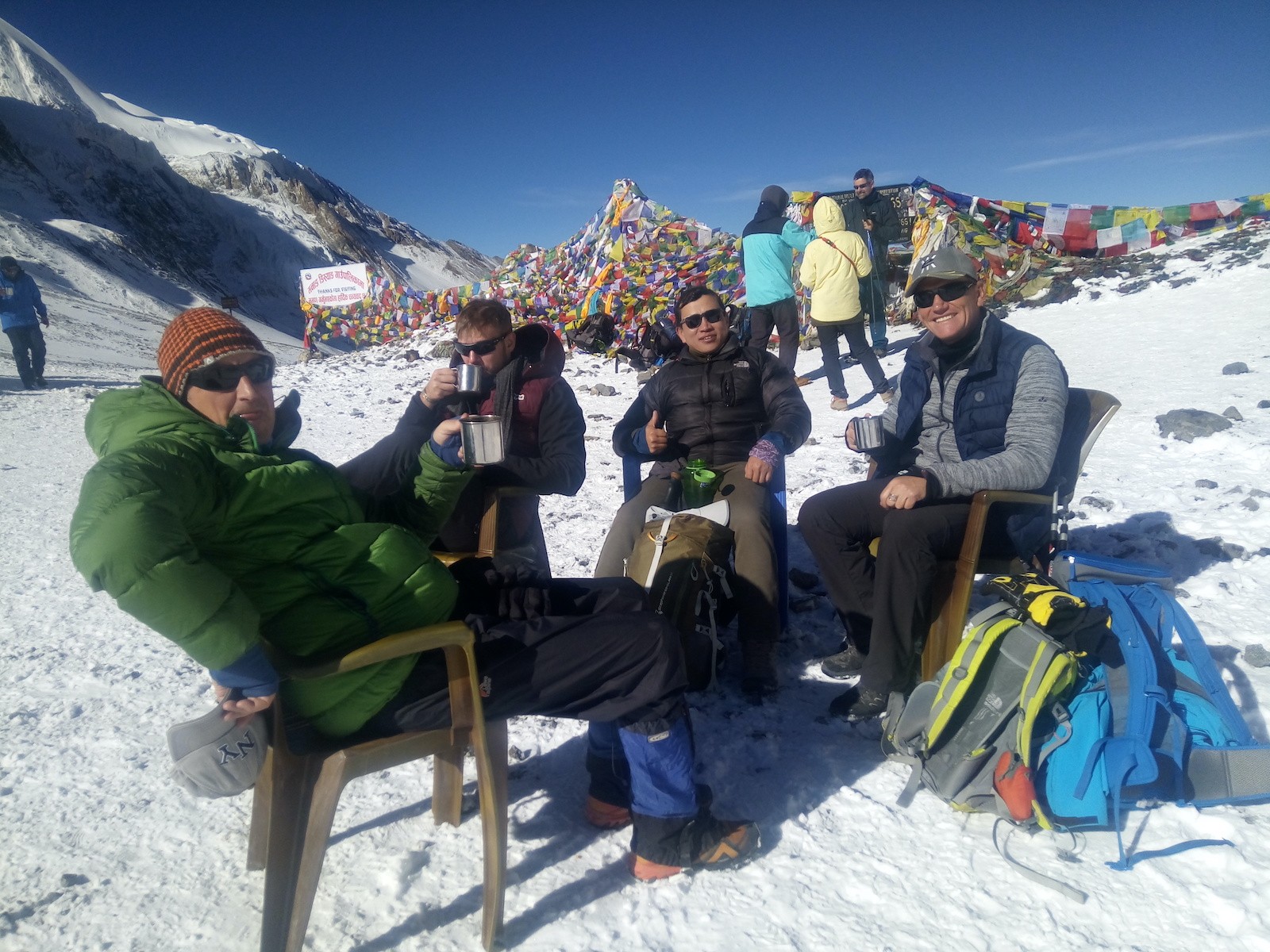 Trekkers celebrating at Thorong La Pass (5,416 m) in October, enjoying hot tea amid colorful prayer flags, light snow, and breathtaking Himalayan peaks under a clear blue sky.