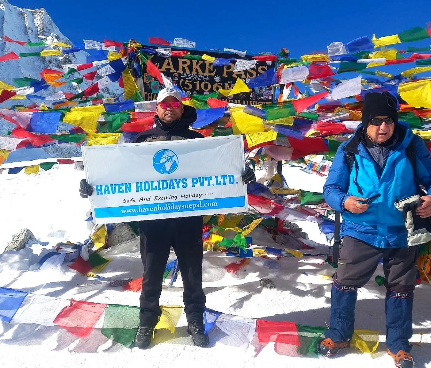 Trekkers crossing Larke Pass surrounded by snow-covered terrain, colorful prayer flags, and panoramic Himalayan peaks on the Manaslu Circuit Trek