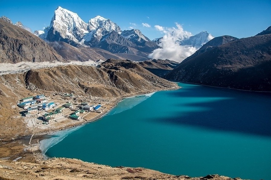 The turquoise waters of Gokyo Lake shimmer beneath towering snow-covered Himalayan peaks, with the village of Gokyo nestled along the shoreline under clear September skies in Nepal's Everest region.