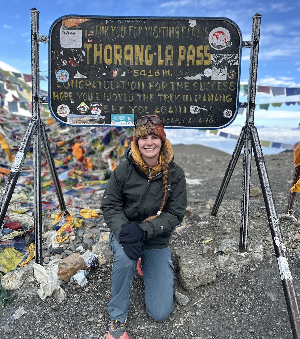 Trekkers  at Thorong La Pass surrounded by colorful prayer flags, snow-covered terrain, and spectacular views of the Himalayan mountain range at 5,416 meters above sea level.