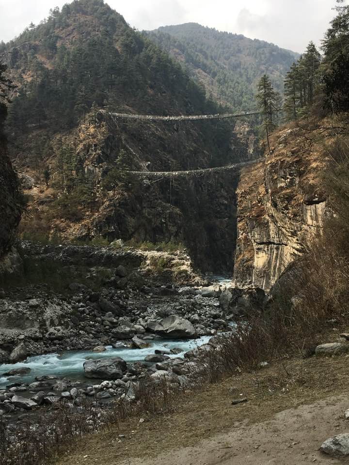 A dramatic view of the dual high-suspension Hillary Bridges draped in colorful prayer flags spanning a deep, rocky canyon over the rushing Dudh Koshi River, framed by steep pine-forested cliffs in the Himalayas. 