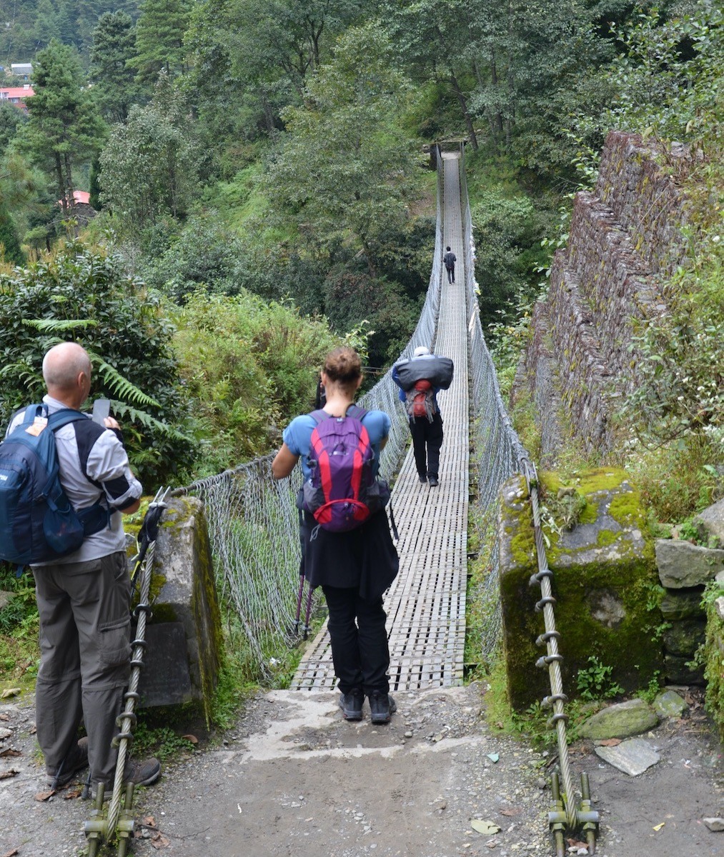 Trekkers crossing a suspension bridge through lush green forests in Nepal's lower valleys, enjoying sunny daytime weather on a scenic Himalayan trekking trail.