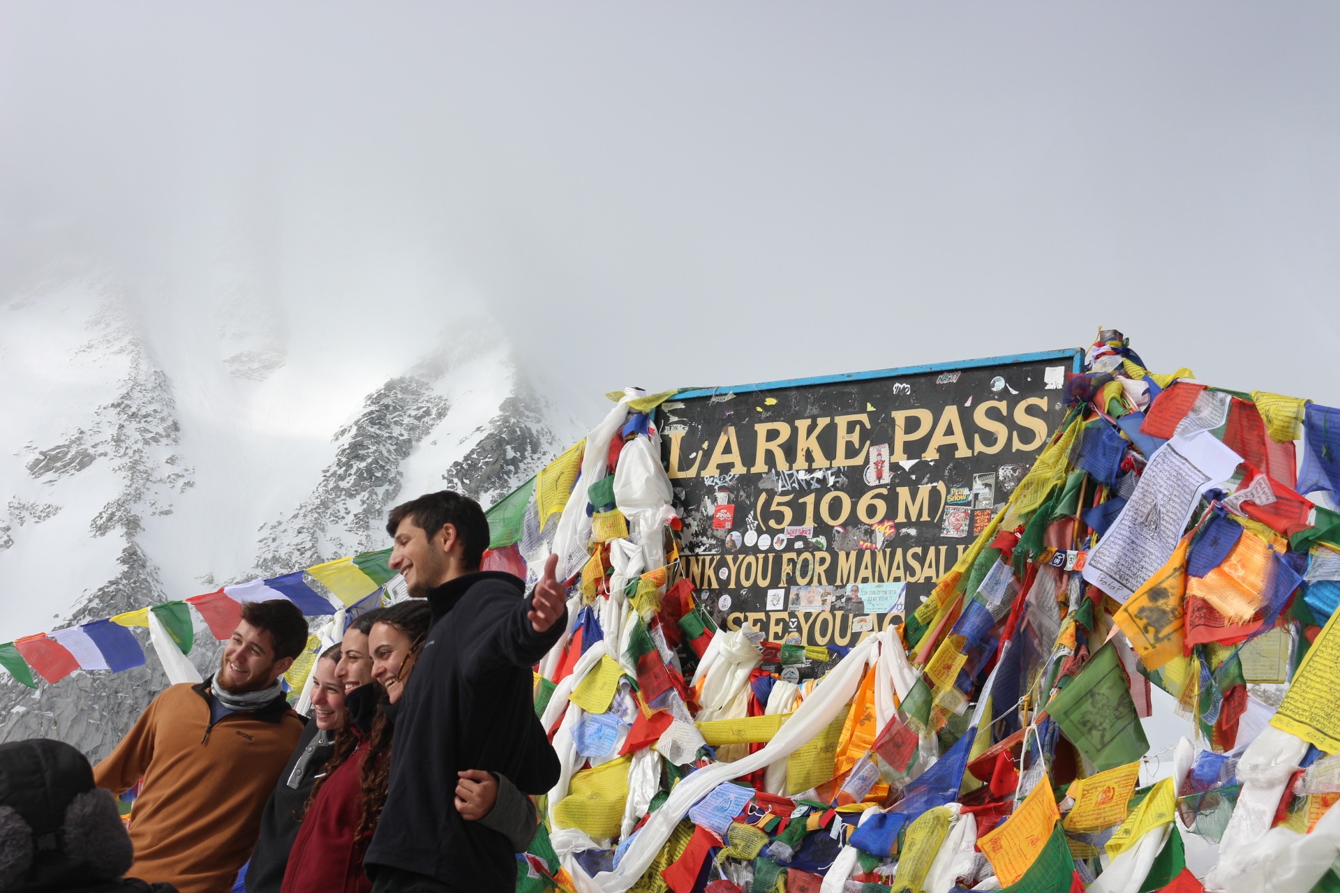 Trekkers reaching the summit of Larke Pass surrounded by snow-covered Himalayan peaks, prayer flags, and high-altitude alpine terrain.