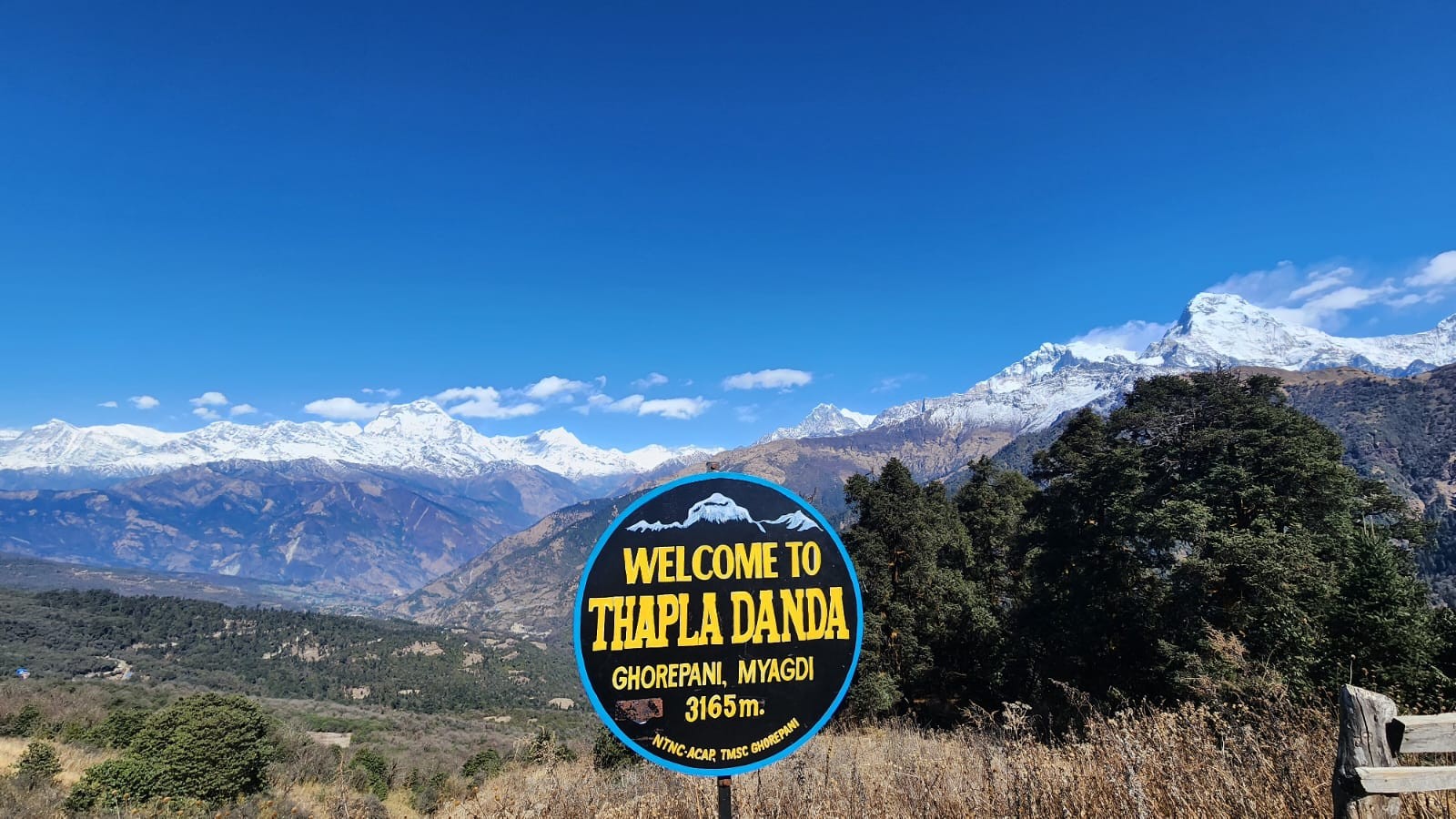 Welcome to Thapla Danda (3,165 m) sign in Ghorepani, Myagdi, with spectacular panoramic views of the Annapurna and Dhaulagiri mountain ranges beneath a clear blue Himalayan sky.