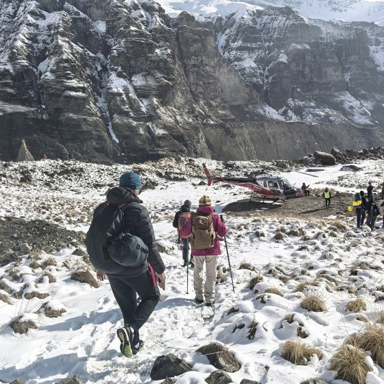 Trekkers walking through fresh snow near Annapurna Base Camp as a helicopter lands, surrounded by towering cliffs and snow-covered Himalayan mountains.