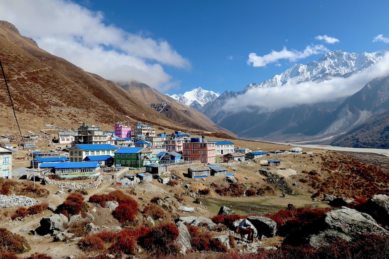 Kyanjin Gompa surrounded by towering snow-capped Himalayan peaks, featuring the historic monastery, traditional stone lodges, prayer flags, and breathtaking mountain scenery.