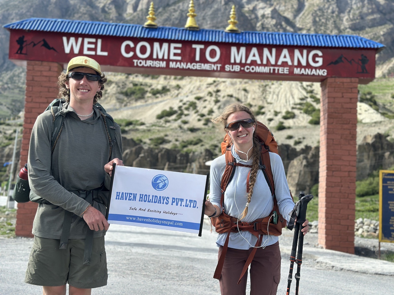Trekkers wearing lightweight, layered hiking clothing and carrying backpacks while walking through Manang Village in daylight, surrounded by clear skies and spectacular Himalayan mountain scenery.