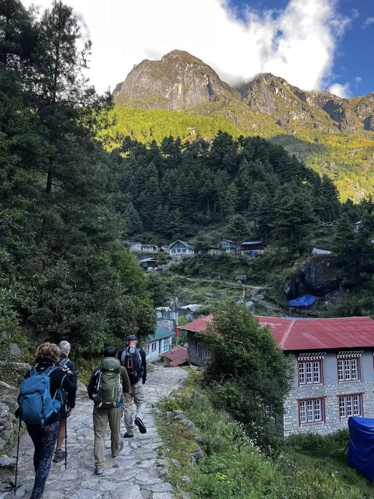 Trekkers walking along a stone-paved trail through a traditional Himalayan village surrounded by green forests and terraced hillsides during Nepal’s monsoon season.