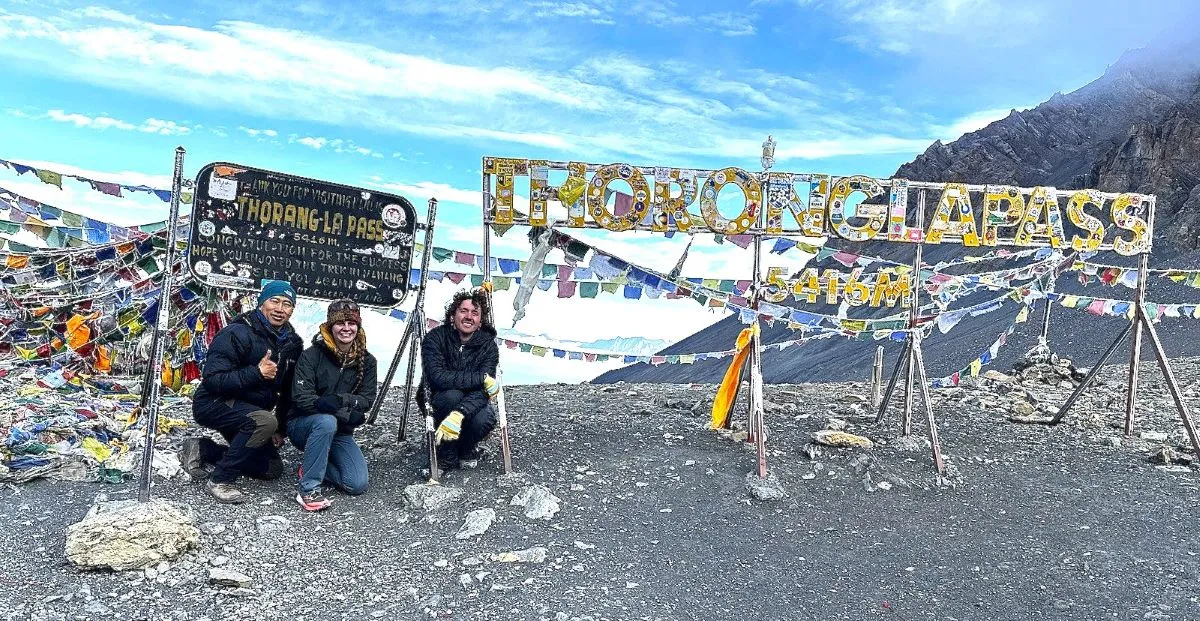 Trekkers celebrating at the summit of Thorong La Pass beside colorful prayer flags and the iconic pass sign, surrounded by snow-covered Himalayan peaks and high-altitude alpine terrain.